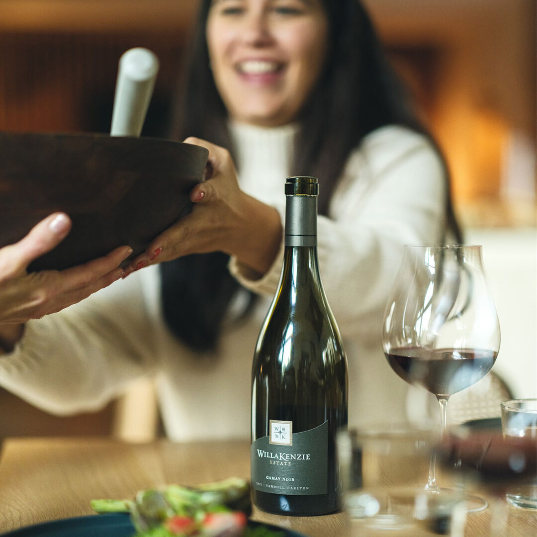 Bottle of Gamay Noir on table next to a woman passing a prepared dish..